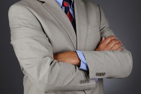Closeup of a businessman wearing a light gray suit standing with his arms crossed. Horizontal format over a light ot dark gray background. Man is unrecognizable.